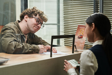 Caucasian young adult man filling out paperwork at counter while young adult woman assisting from opposite side, both interacting in office or service environment