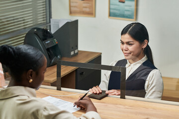 Young adult Hispanic woman sitting behind counter assisting young adult Black woman filling out paperwork at service desk, both women engaged in customer service interaction