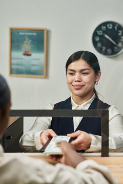Young adult woman working as bank teller handing cash to unseen customer across counter, smiling while performing transaction, clock and framed poster visible in background