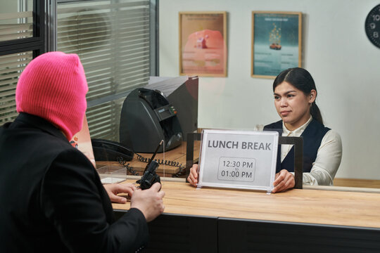 Young adult Caucasian man wearing pink ski mask holding gun confronting young adult teller sitting behind counter with lunch break sign displayed