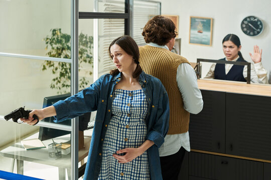Pregnant Caucasian young adult woman holding handgun during bank robbery while Caucasian man standing at counter and young adult woman raising hands behind desk