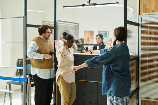 Caucasian man and woman holding guns confronting Black woman and Hispanic woman with hands raised in bank office setting, displaying tense expressions during standoff