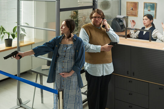 Caucasian young pregnant woman and man holding handguns during bank robbery, man talking on phone, bank teller standing behind counter with hands raised