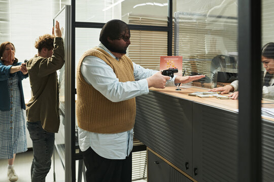 Young man wearing mask robbing bank counter with handgun while teller handing over cash, and young Caucasian man standing with hands raised while woman in mask pointing gun in background
