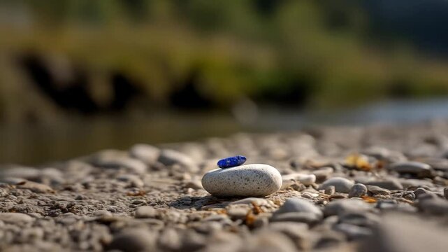 Small blue stone balances atop a larger, grey stone on a stony riverbed, soft focus background