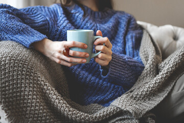 Close up female hands holding blue cup of tea or coffee. Woman in knitted sweater and cozy plaid relaxing at home on sofa, warm and peaceful indoor atmosphere
