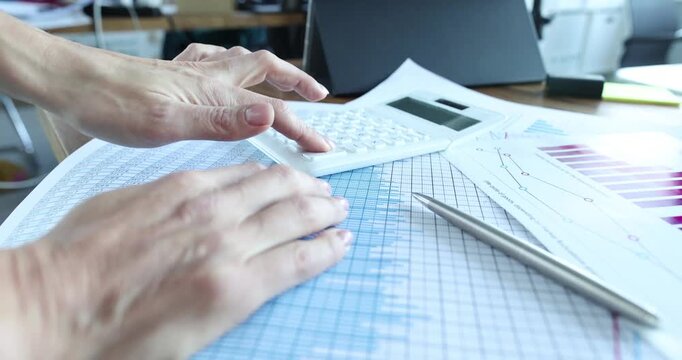Accountant hand presses calculator buttons on finance charts near spreadsheet. Worker verifies totals and enters figures preparing monthly report