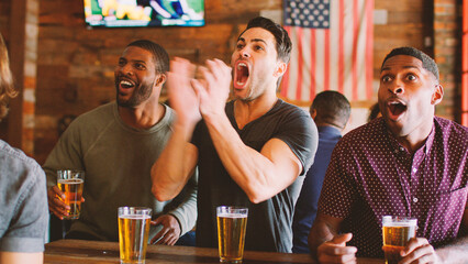 Group Of Male Friends Celebrating Whilst Watching Game On Screen In Sports Bar