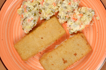 An overhead close-up of two golden-brown breaded fish filets and a creamy portion of French salad (Croatian way) on an orange-rimmed plate