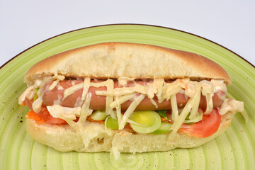 A juicy hot dog in a soft bun, fully loaded with shredded cheese, creamy sauce, sliced tomatoes, and chopped leeks, served on a green striped plate