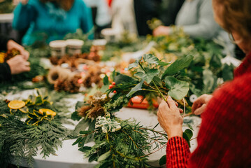 A group of people are making wreaths with green leaves and pine cones. Scene is festive and joyful, as the people are working together to create beautiful decorations for the holiday season