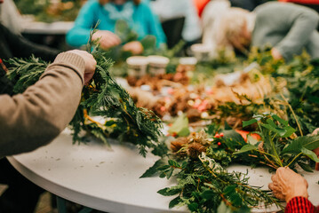 A group of people are gathered around a table, working on a wreath. The wreath is made of green leaves and branches, and the people are using scissors to cut and arrange the materials