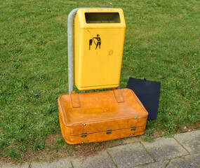 brown leather suitcase lies outside in front of a yellow trash bin on a lawn