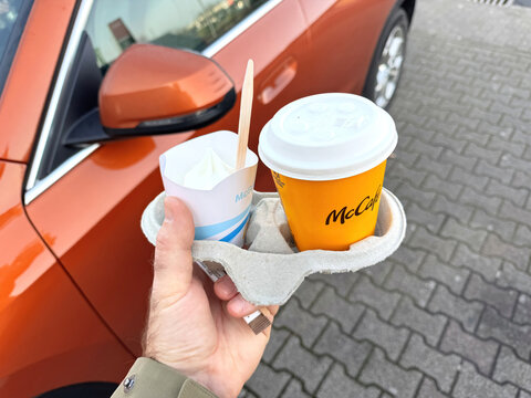 Frankfurt, Germany - Apr 1, 2025: Man holding a McCafe coffee cup and McFlurry ice cream in a cardboard tray near a Tahoe orange BMW electric car parked on a paved surface