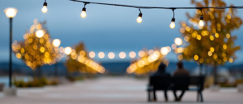 Blurred evening scene with string lights and illuminated trees lining a walkway where two people sit on a bench under a cloudy sky