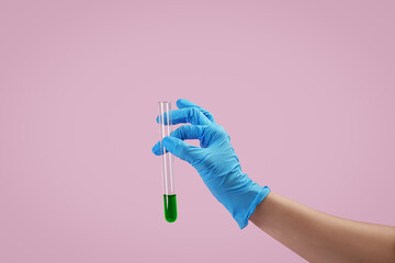 A gloved hand holds a clear test tube containing vibrant green liquid. The background is pink, emphasizing the scientific experiment being conducted, showcasing chemistry.