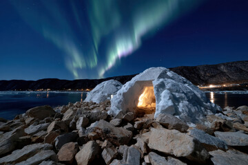 An inviting igloo, illuminated from within, sits on a stony beach while the mesmerizing Northern Lights dance across the night sky, creating a magical winter scene.