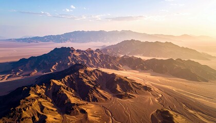 An aerial perspective reveals dramatic, rugged desert mountains in the foreground, transitioning to softer, layered mountain ranges and flat plains under a warm