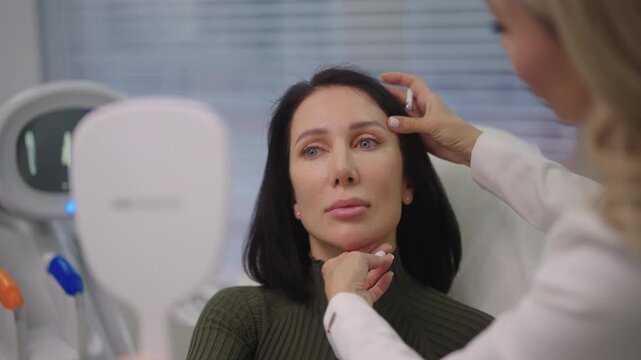 Portrait of middle-aged female client consulting with medical aesthetic specialist who carefully marks facial contours and treatment areas with white pencil during pre-procedure planning in clinic.