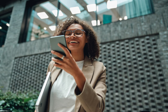 woman checking smartphone standing in atrium creating social content, smiling and composing caption, modern