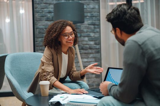 smiling consultant advising client in modern office, relaxed coaching session with laptop, documents, coffee