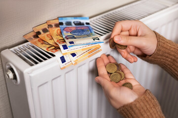 A girl in a sweater is counting money by the radiator. The room is cold, so the heating is needed.