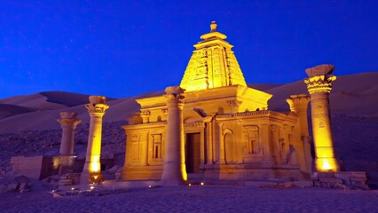 A stone temple with Egyptian-style architecture stands illuminated in a desert landscape at night, with sand dunes and a starry sky in the background.