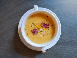 A white bowl of creamy yellow soup topped with corn, small meat slices, herbs, and grated cheese, placed on a gray table.