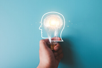 Creative idea and innovation concept. Hand holding lightbulb with glowing brain in head silhouette. Brainstorming, AI, knowledge, and new solution on blue background with copy space.
