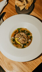 A white plate with a dish of food on it. The plate is on a wooden table. The food on the plate is a soup with vegetables