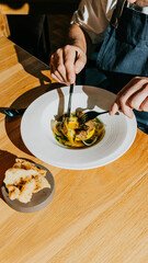 A man is eating a meal with a fork and knife on a wooden table. The meal consists of a white plate with a piece of bread and a bowl of food. The man is wearing an apron and he is enjoying his meal