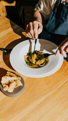 A man is eating a meal with a fork and knife. The meal is served in a white bowl and includes a piece of bread. The man is sitting at a wooden table