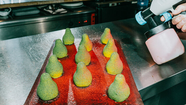 A person is spraying a tray of green pear-shaped pastries with a mist. The tray is filled with pastries and the mist is coming out of a spray bottle - Powered by Adobe
