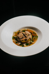 A white plate with a brownish food item on it. The food item is surrounded by green leaves and has a brown crust. The plate is placed on a black background