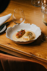 A plate of food is on a wooden table with a chair in front of it. The table is set with a wine glass, a fork, and a knife. The food on the plate is a fish dish with a creamy sauce