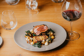A plate of meat and vegetables sits on a wooden table next to a wine glass. The wine glass is half full, and the plate is garnished with a few pieces of broccoli. Scene is elegant and sophisticated