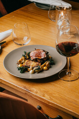 A plate of food with a glass of wine on a table. The plate has meat and vegetables, and the wine glass is filled with red wine. The table setting is elegant and sophisticated, with a knife