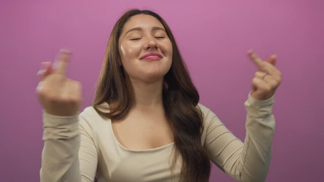 Young hispanic woman wearing beige top raises both middle fingers with playful smirk in pink studio; defiance rebellion independence boldness.