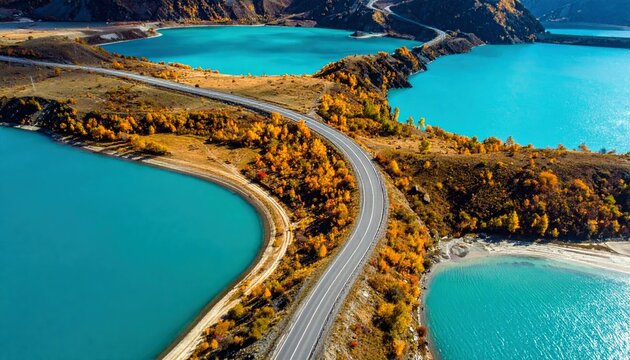 An aerial view captures a winding asphalt road traversing a landscape dotted with trees in autumn colors, bordered by striking turquoise lakes.