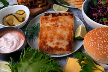 Fishburger ingredients: white fish fillet patty, bun, sauce, pickles, red cabbage and carrot salad, cheddar cheese, french fries, lettuce, and lime wedges on a gray table