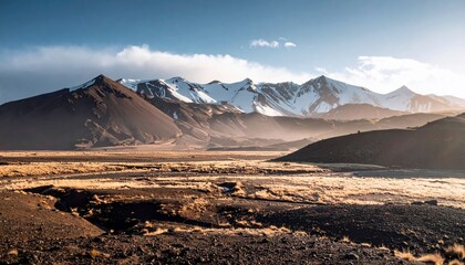 A panoramic view of a rugged mountain range with snow-covered peaks, set against a clear blue sky with scattered clouds, overlooking a dry, grassy valley.