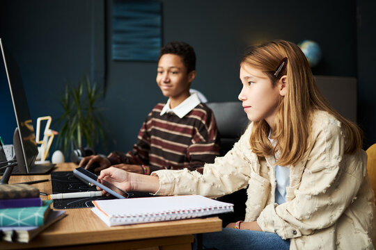 Teenage Caucasian girl using digital tablet at desk while teenage Black boy working on computer in background, both focused on tasks in modern classroom or study environment - Powered by Adobe