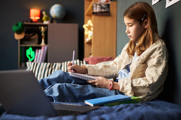 Teenage Caucasian girl sitting on bed writing in notebook with pen, focused on studying with open...