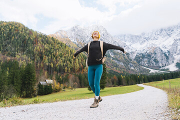 Young attractive woman hiking in nature. Beautiful happy girl stands on road looking at mountains view. Autumn or winter nature landscape. Photo of female having a walk with a backpack outdoors.