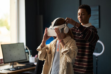 Teenage Black boy helping Caucasian teenage girl put on virtual reality headset in modern room, both standing near desk with computer monitor, engaging in immersive technology experience