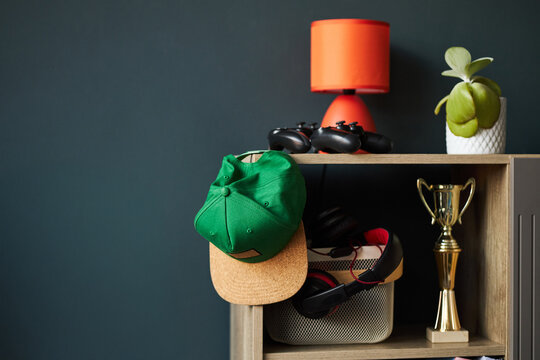 Shelf displaying green baseball cap hanging over edge, two video game controllers, gold trophy, headphones in basket, orange lamp, small potted plant on wooden surface