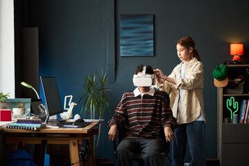 Asian teenage girl adjusting virtual reality headset on seated Asian teenage boy in modern workspace, computer and creative supplies on desk, plant and decor in background