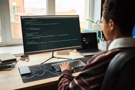 Teenage Black boy sitting at desk using computer monitor and keyboard, coding or programming software, focused on screen, modern workspace with window in background, hands on keyboard