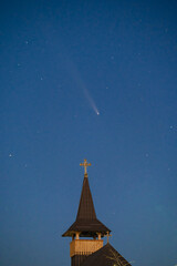 Celestial and Spiritual: Rare Comet ATLAS 2024 Shines Brightly in the Night Sky Behind the Illuminated Cross of an Old Wooden Church, Symbolizing Faith and Science