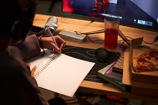 Teenager with headphones sitting at desk writing in notebook while gaming on computer, pizza and drink nearby, focusing on multitasking between studying and playing video game